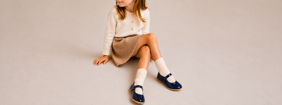 A young girl sits on a bench in a boutique, wearing a navy blue dress, white tights, and black Mary Jane shoes while tying one shoe. A dark navy shoebox sits on the floor next to her.