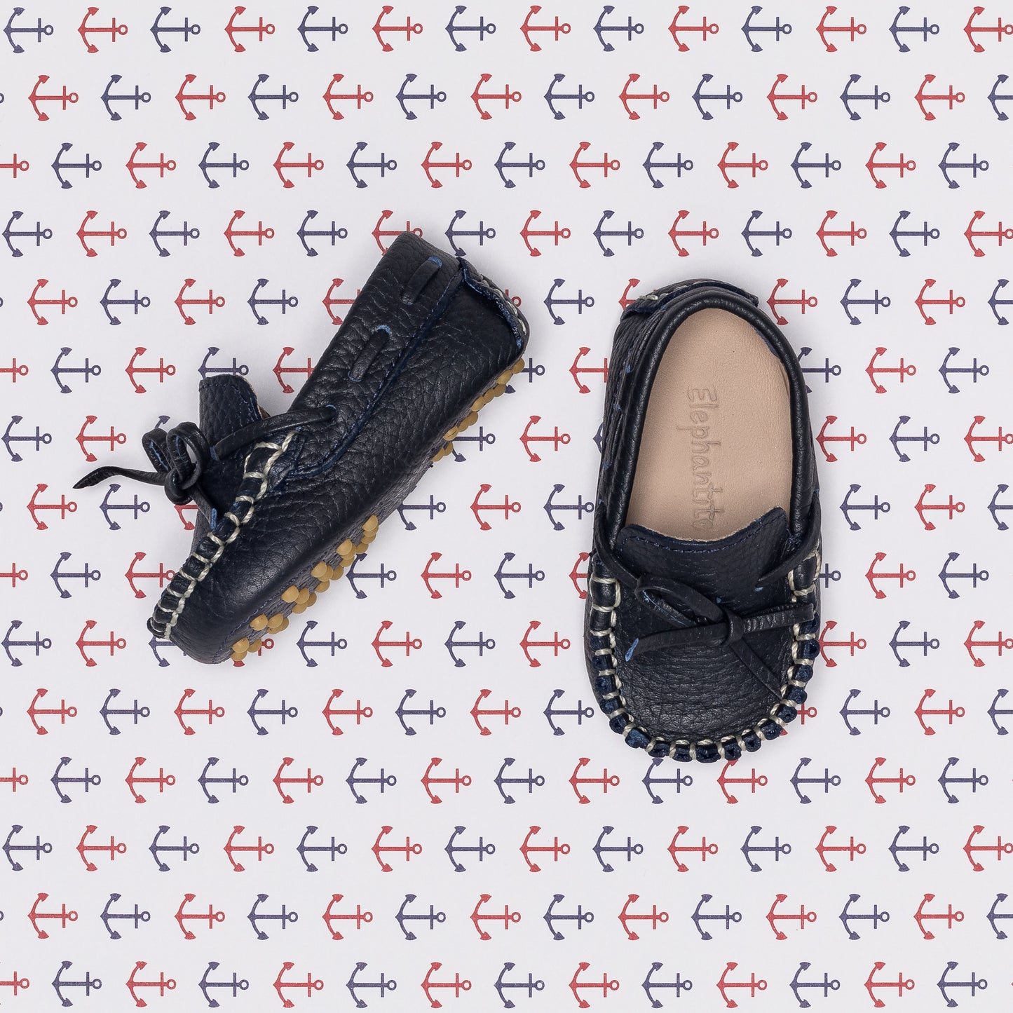 Pair of navy blue baby driver loafers with bow detail and textured soles on anchor-patterned background.