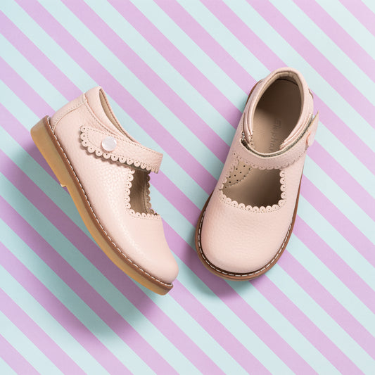 Light pink leather girls’ Mary Jane shoes with scalloped strap and tan soles on striped pastel background.