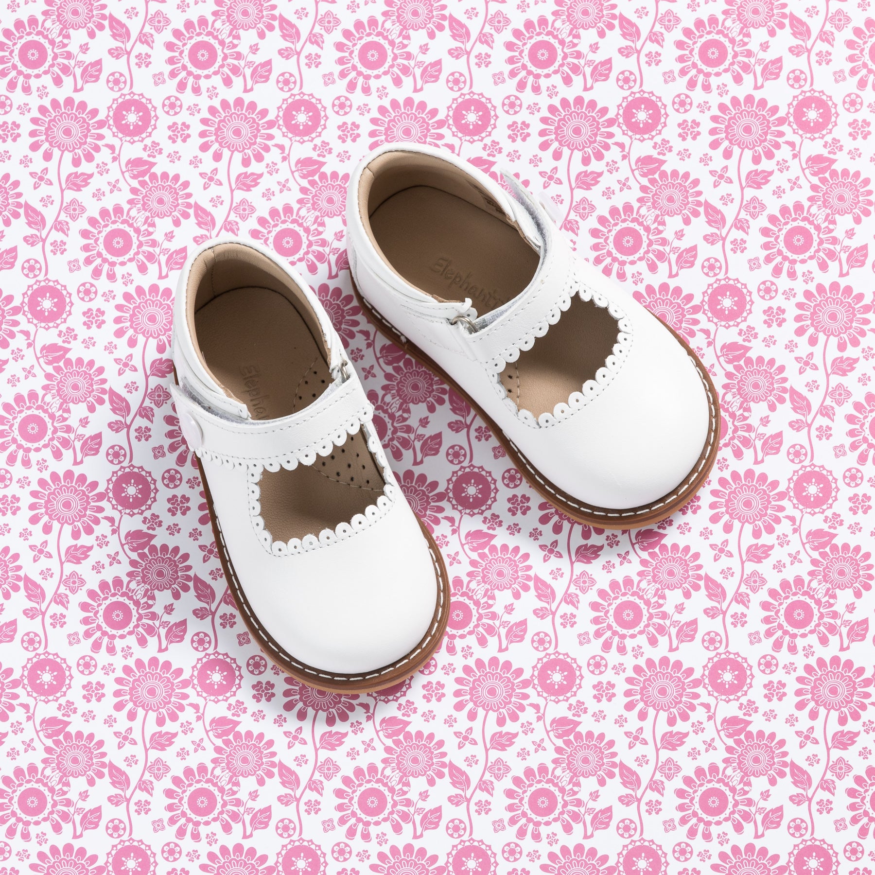 White leather girls' Mary Jane shoes with scalloped edges and brown rubber soles on pink floral background.