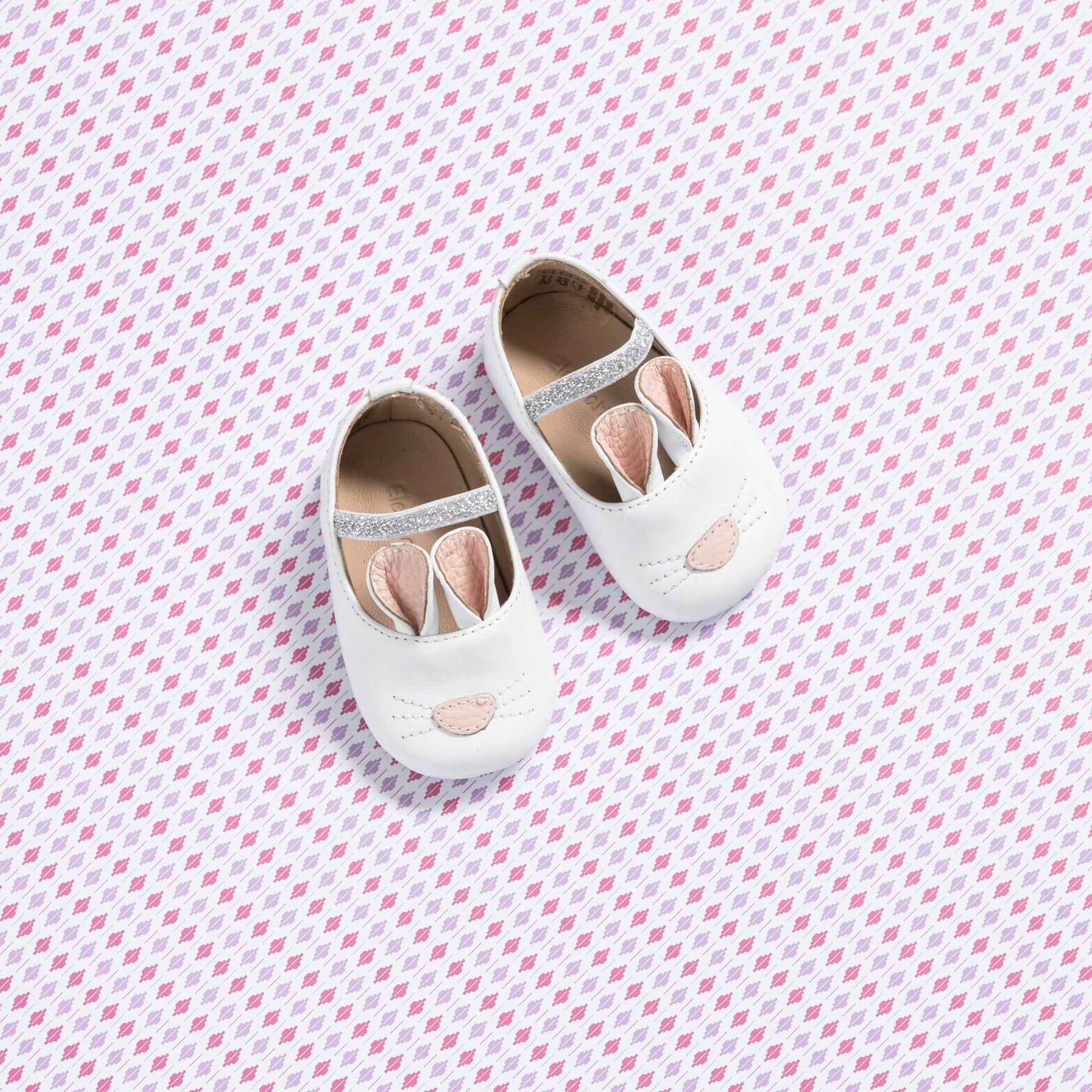 White baby slippers shaped like bunnies with pink accents and soft soles on a patterned background.