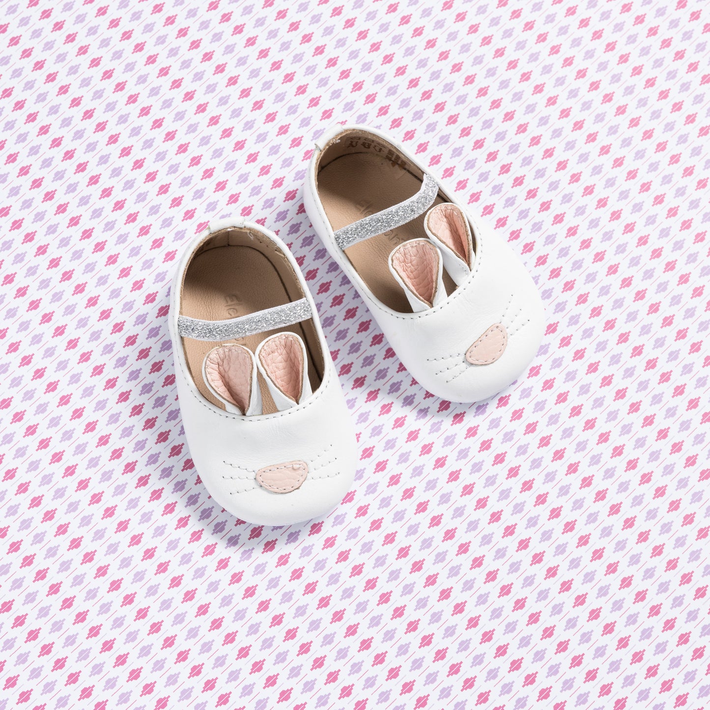 White baby slippers with bunny ears and embroidered nose detail on soft fabric background.