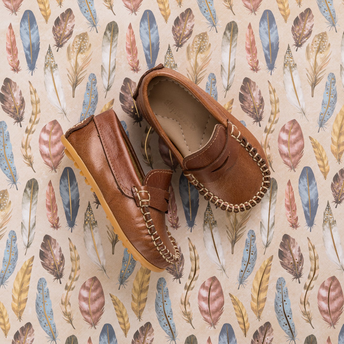 Natural brown toddler moccasin shoes with stitched detailing and soft sole on feather-patterned background.