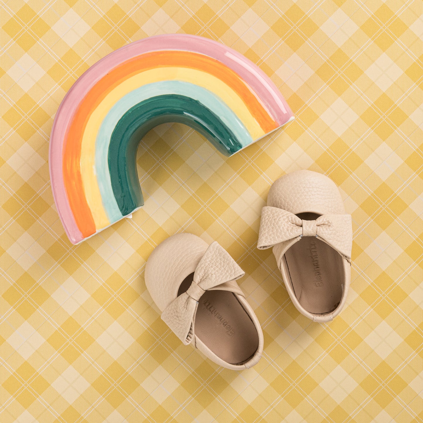 Cream-colored baby ballerina shoes with textured bows on a yellow checkered background, paired with a ceramic rainbow.