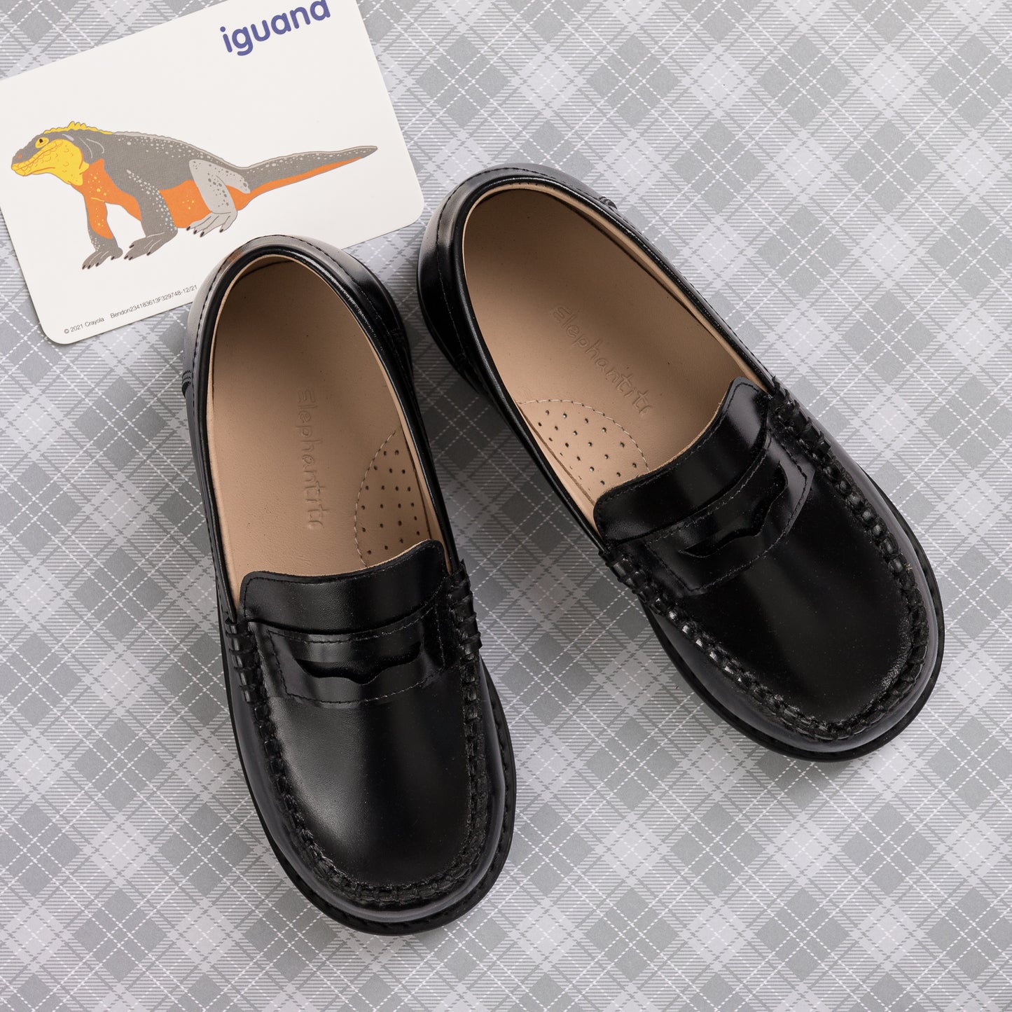 Black leather boys' moccasins with a penny strap and cushioned perforated insole on a gray checkered background.