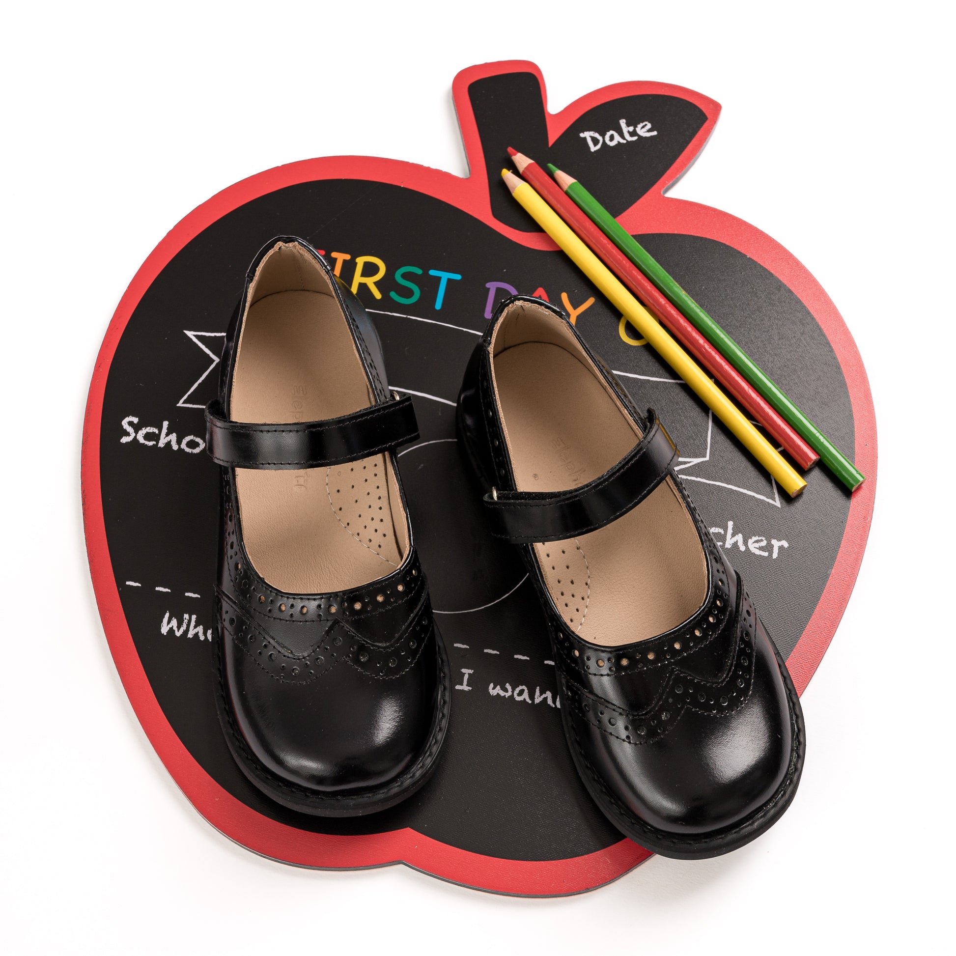 Black leather Mary Jane shoes with perforated detailing and adjustable strap on a red apple-shaped chalkboard with colorful.