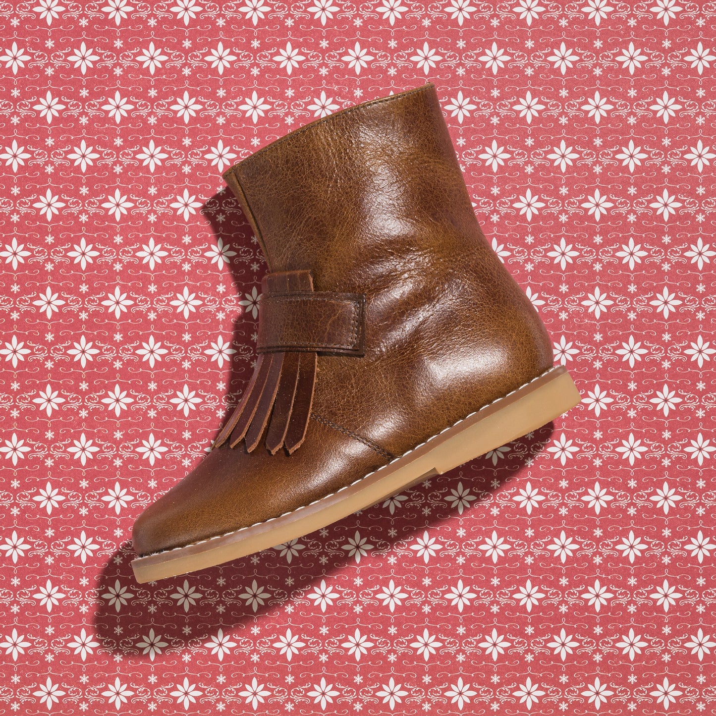 Brown leather toddler bootie with side fringe and tan rubber sole on patterned background.