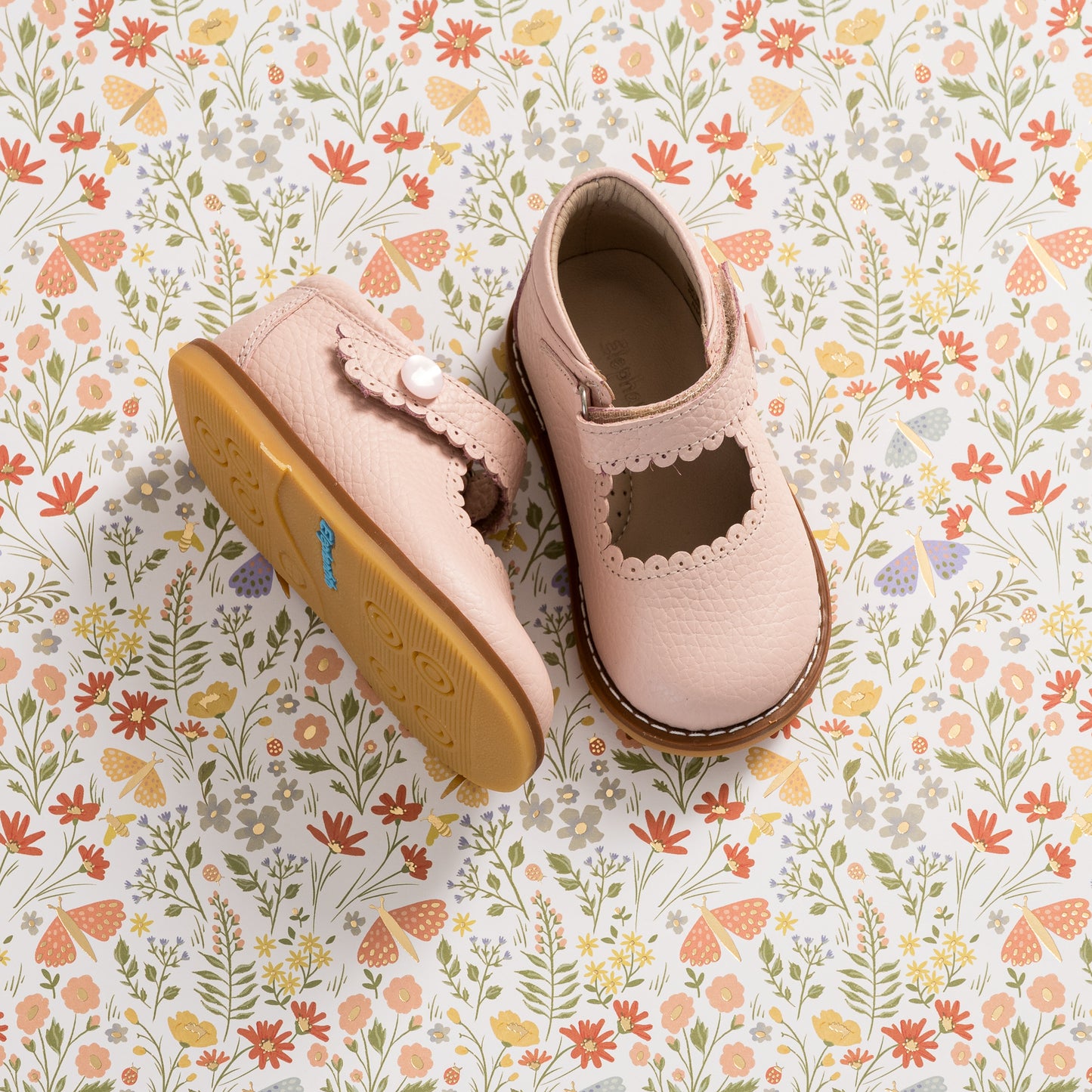 Pink leather toddler Mary Jane shoes with scalloped strap and rubber sole on floral background.