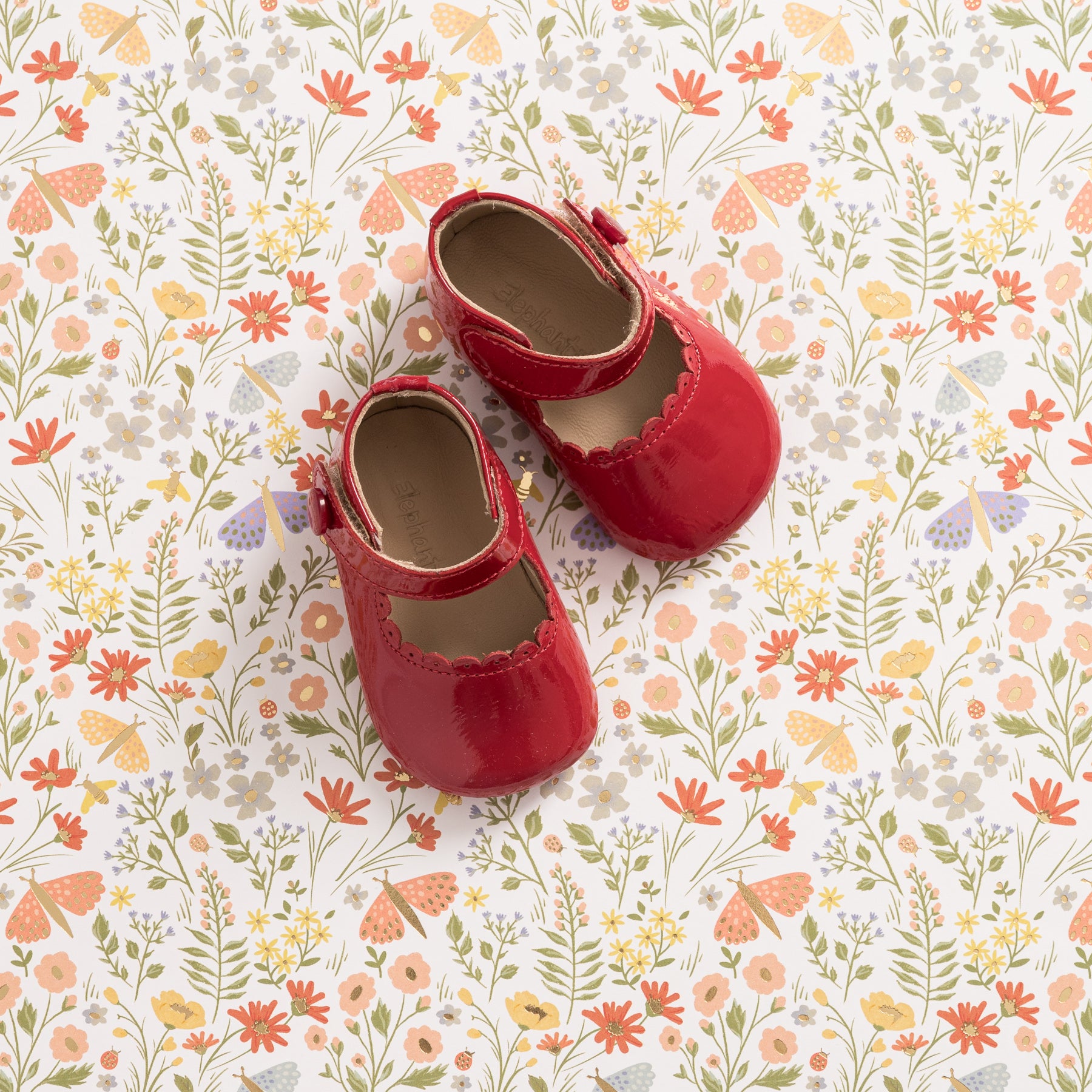 Red patent leather baby Mary Jane shoes with scalloped edges on floral background.