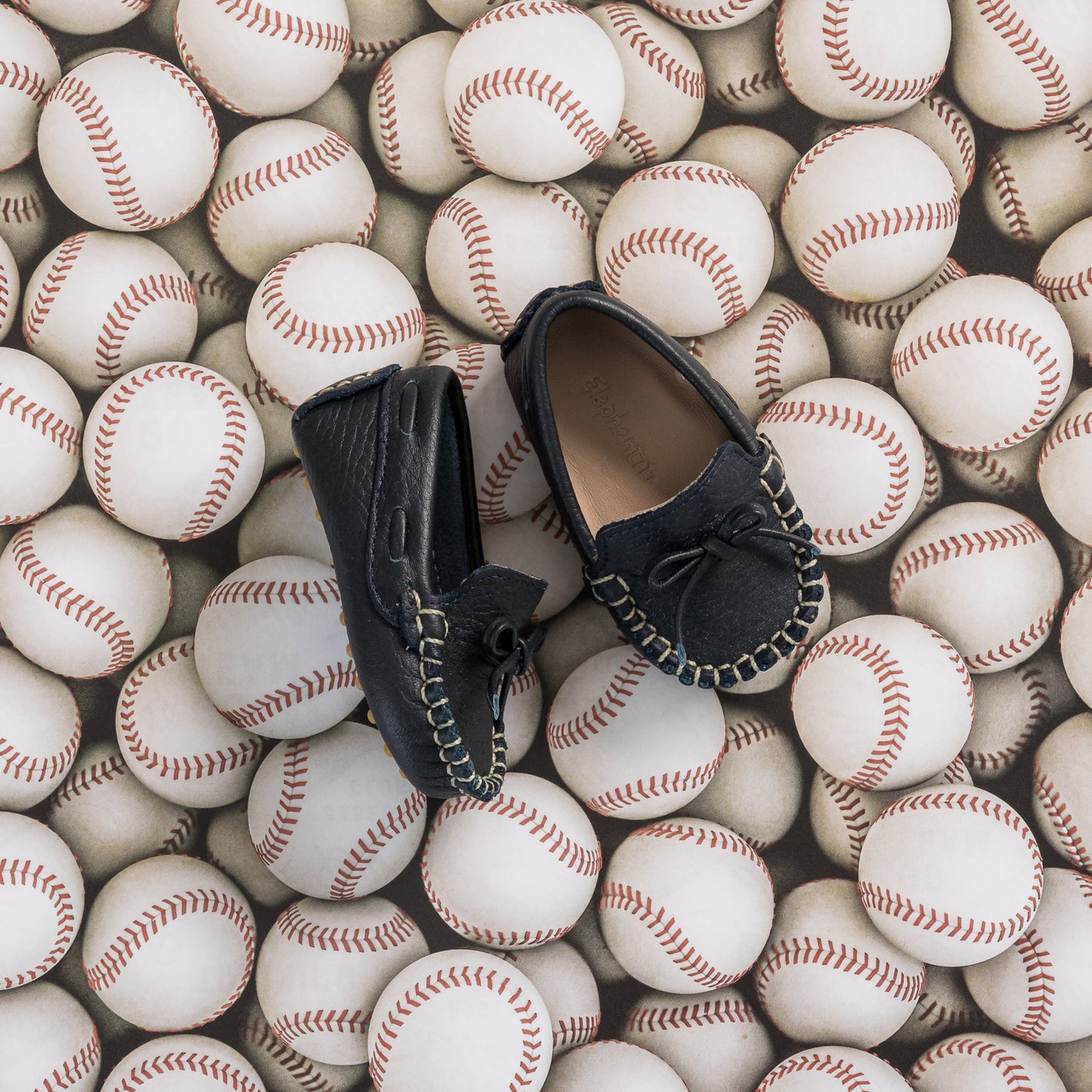 Navy blue baby driver loafers with black bow and stitching details on baseball background.