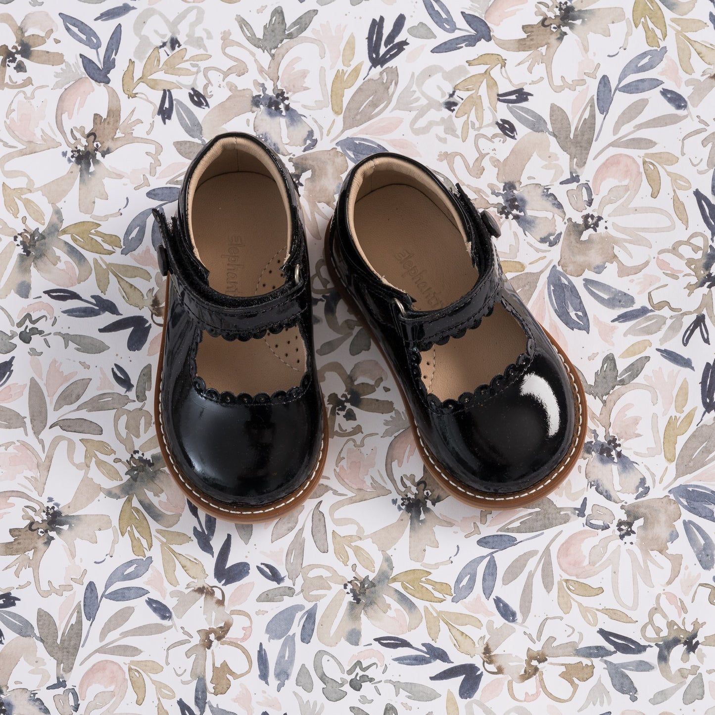 Glossy black patent leather Mary Jane toddler shoes with scalloped edges and brown soles on floral background.