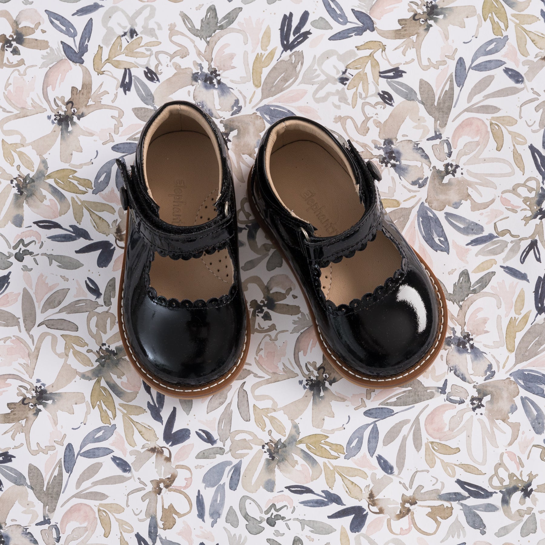 Glossy black patent leather Mary Jane toddler shoes with scalloped edges and brown soles on floral background.