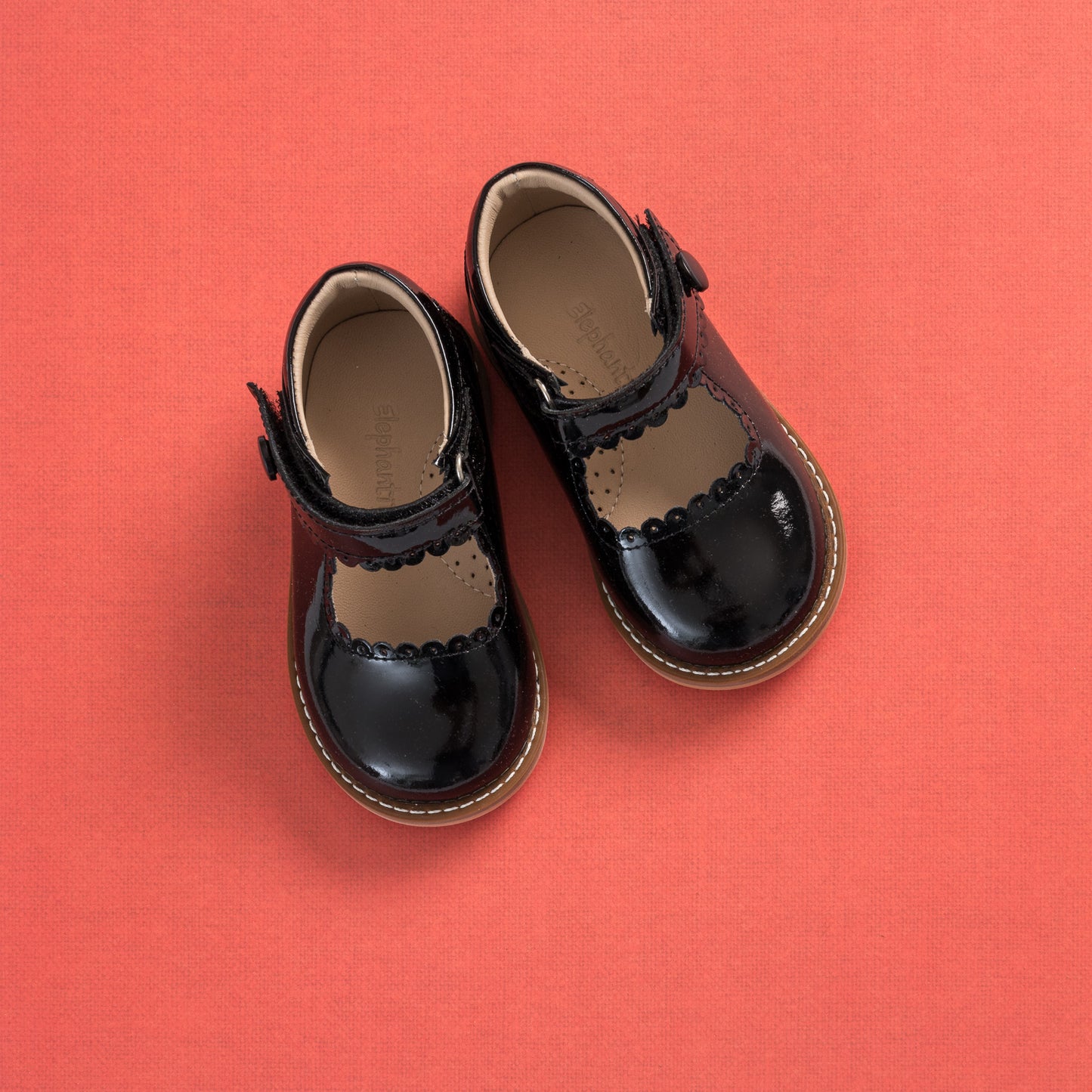 Black patent leather toddler Mary Jane shoes with scalloped edges and brown stitched soles on coral background.