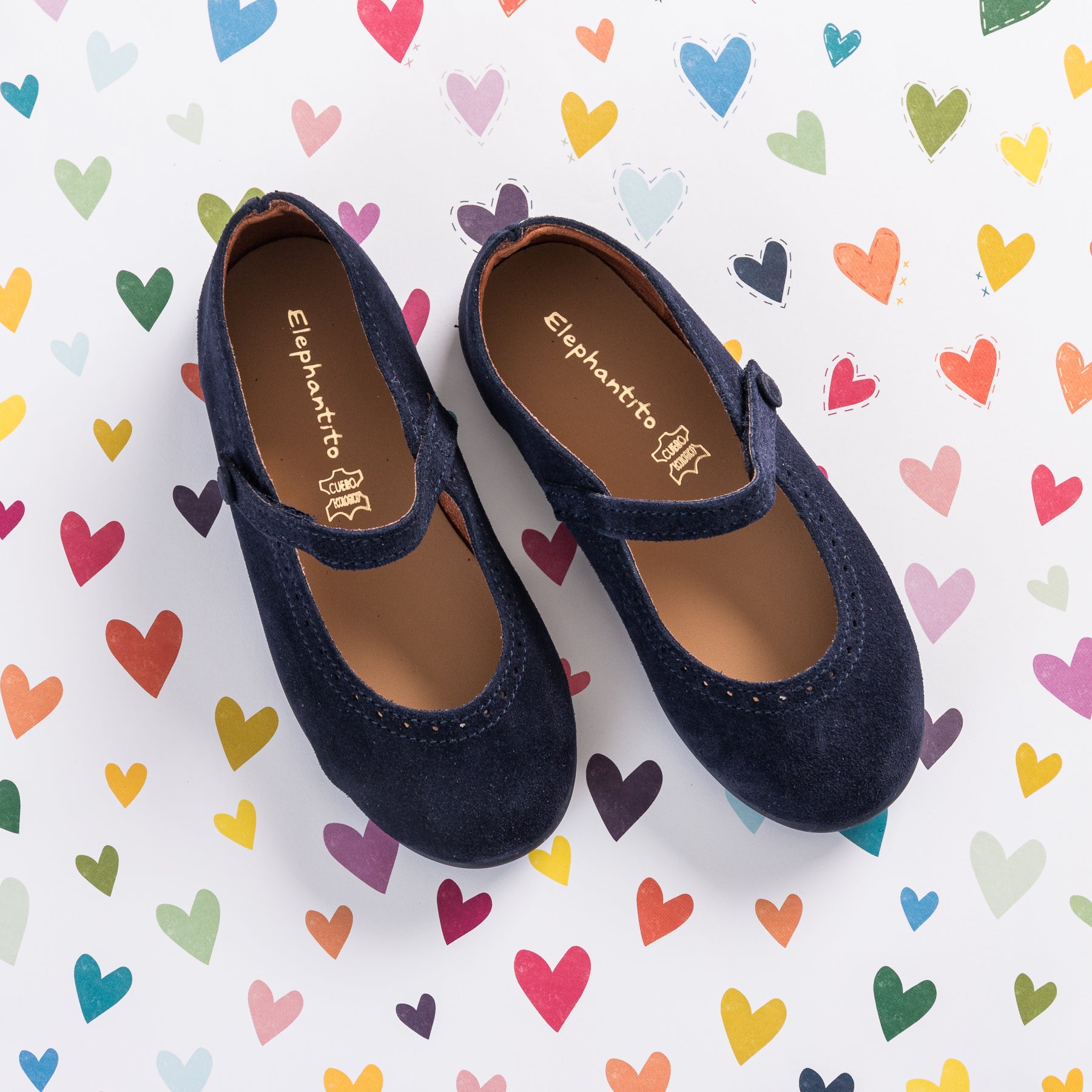 Navy blue suede Mary Jane shoes for toddlers with stitched detailing and a velcro strap on a heart-patterned background.