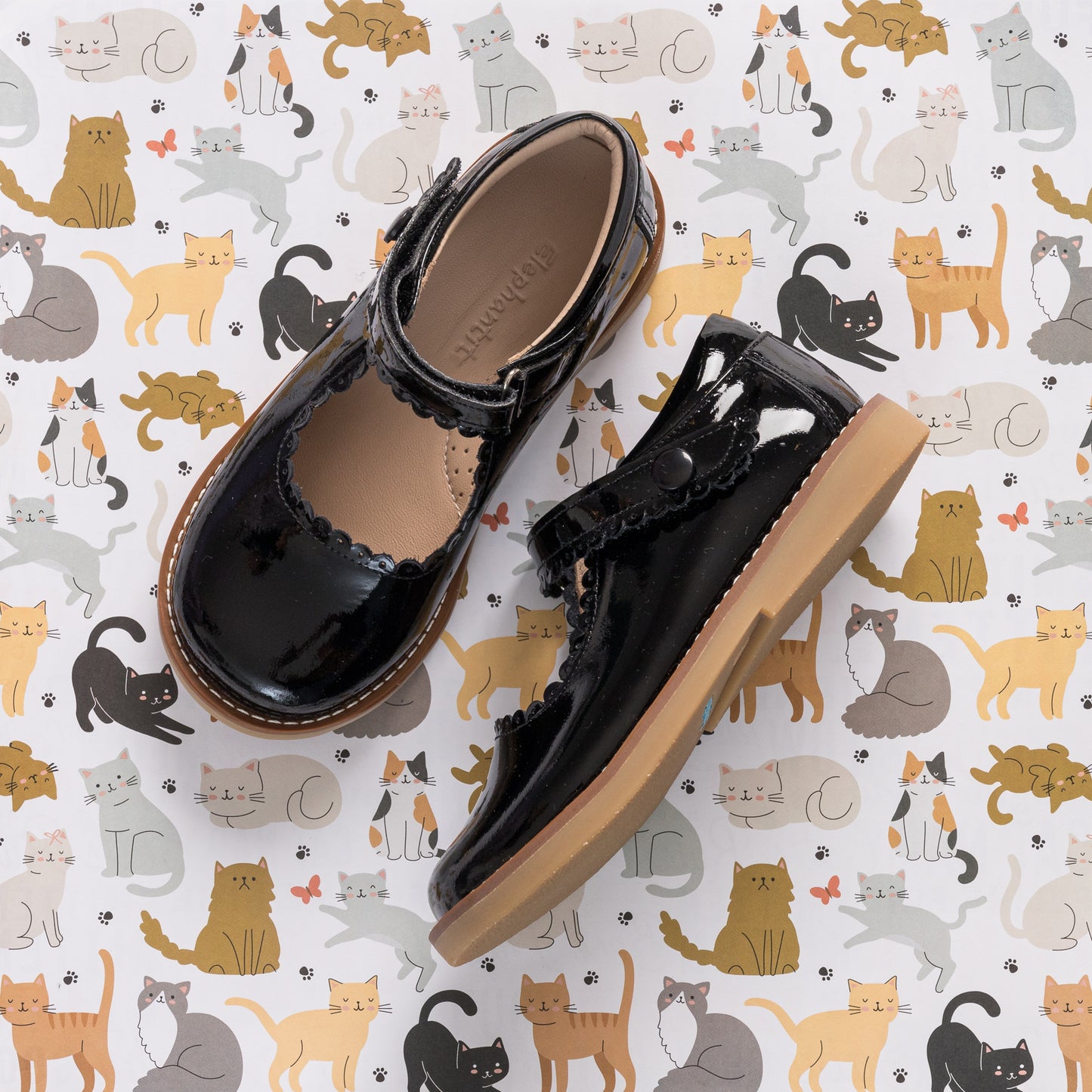 Black patent leather Mary Jane shoes with scalloped edges and buckle strap on a cat-patterned background.