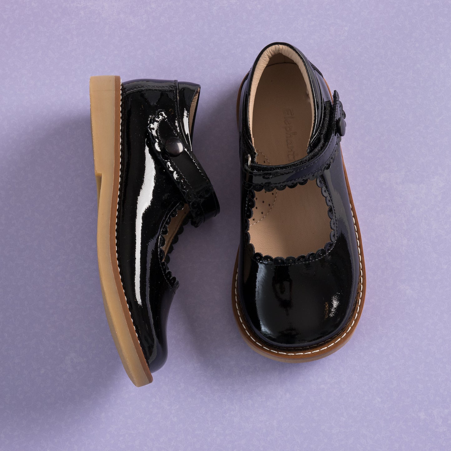 Black patent leather girls Mary Jane shoes with scalloped edges and tan rubber soles on purple background.