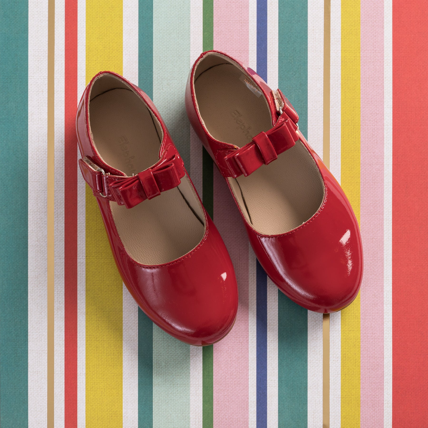 Red patent leather Mary Jane shoes for girls with a velcro bow strap on colorful striped background.