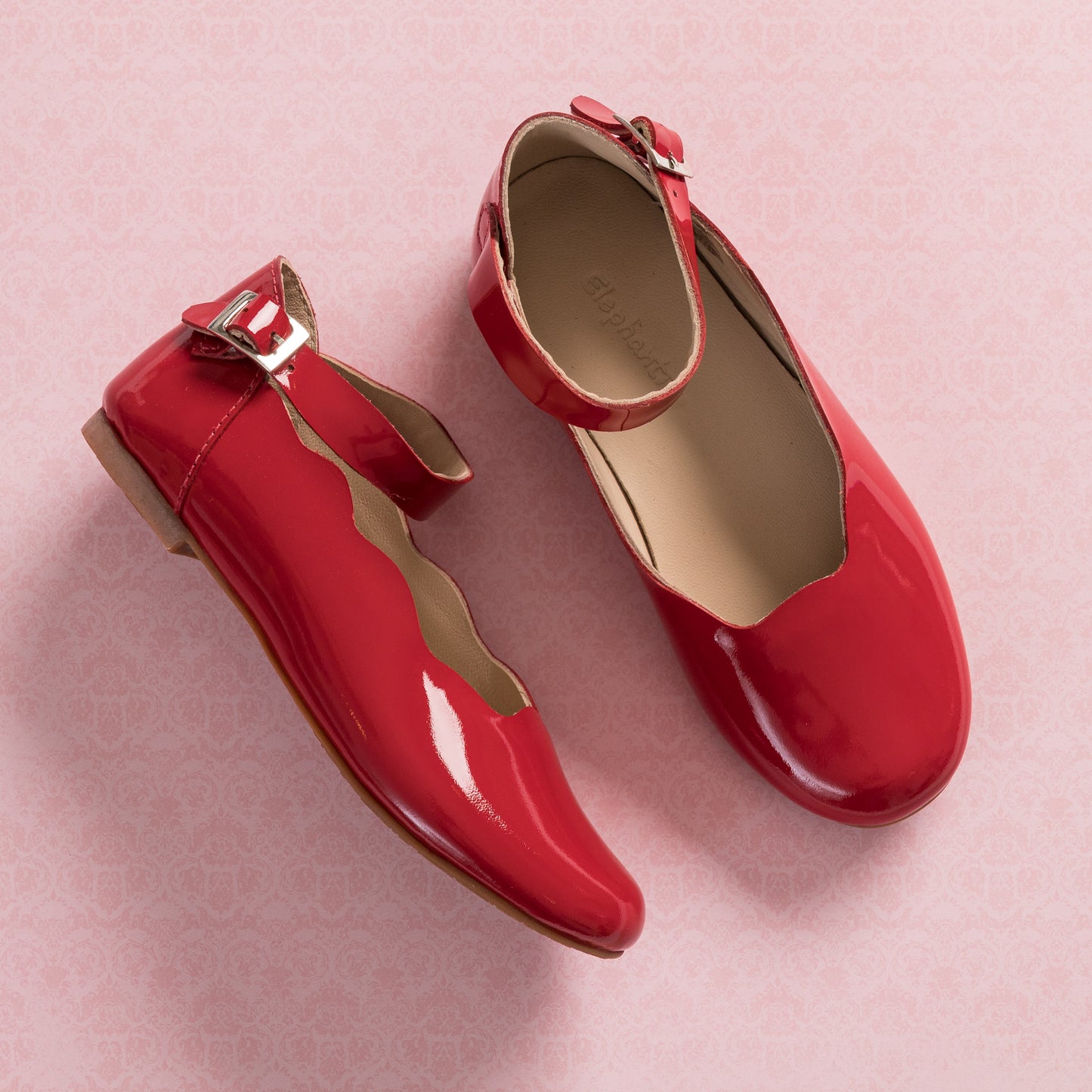 Red patent leather children's flats with scalloped edges and ankle buckle straps on pink background.