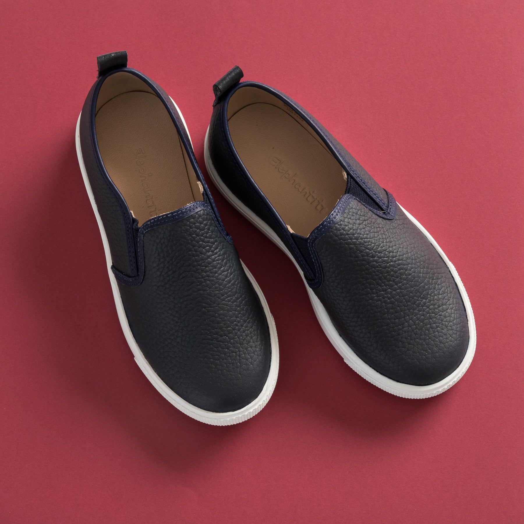 Navy blue textured slip-on toddler shoes with white soles and elastic side panels on red background.