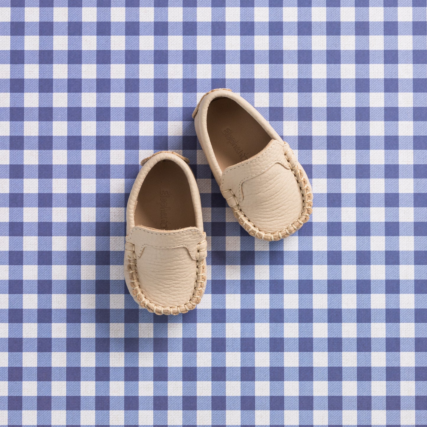 Cream leather baby moccasins with stitched detailing on blue and white checkered surface.