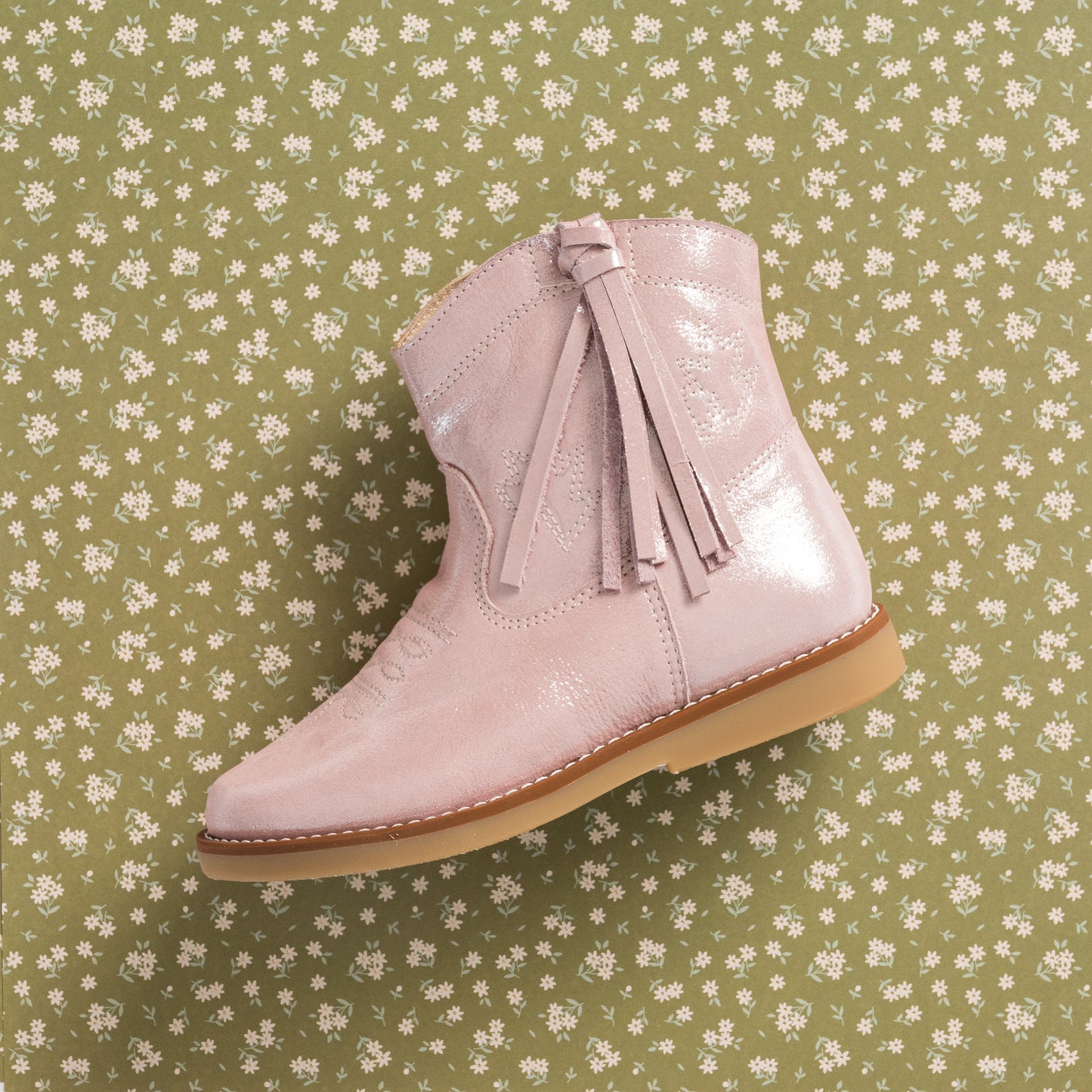 Metallic pink suede toddler boot with side tassels and rubber sole on floral background.