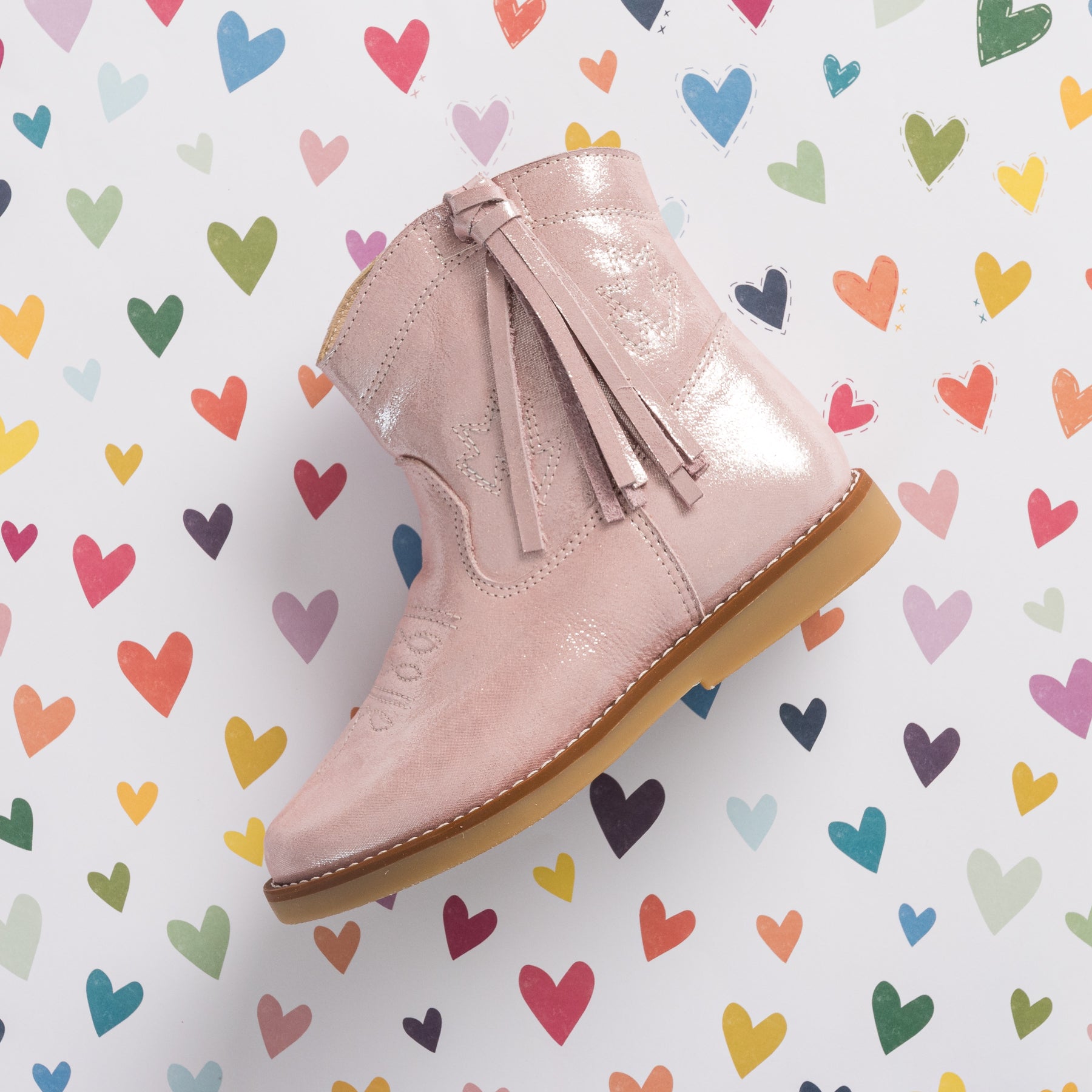 Metallic pink suede toddler boot with tassel zipper and light brown sole against colorful heart background.