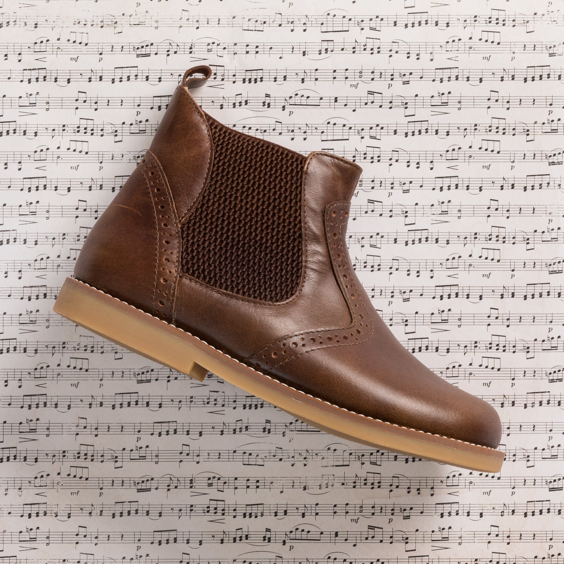 Brown leather toddler bootie with elastic side panels and tan rubber sole on sheet music background.