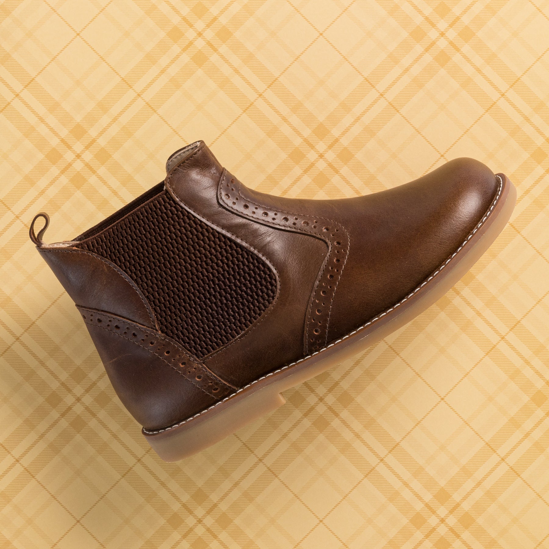 Brown leather toddler bootie with elastic side panels and pull tab on plaid background.