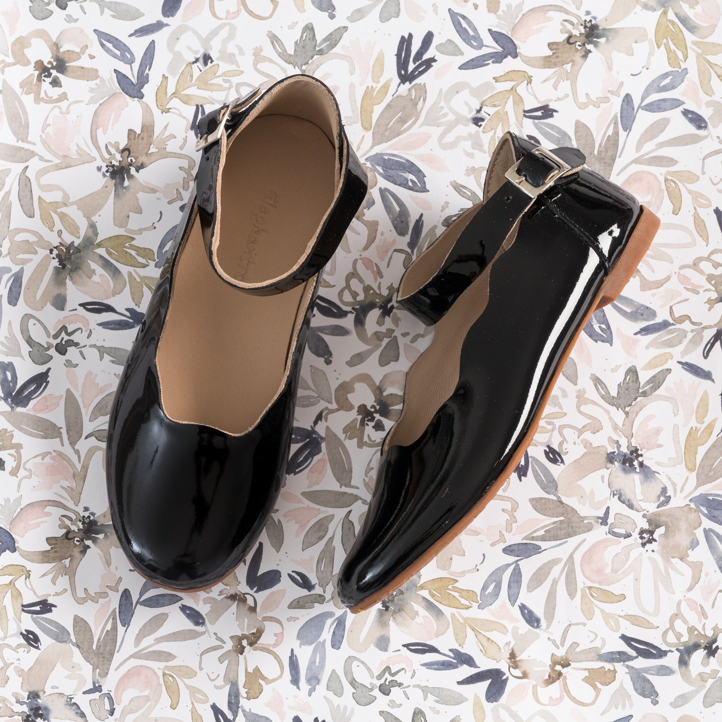 Black patent leather girls' ballet flats with ankle straps and buckle closures on floral background.