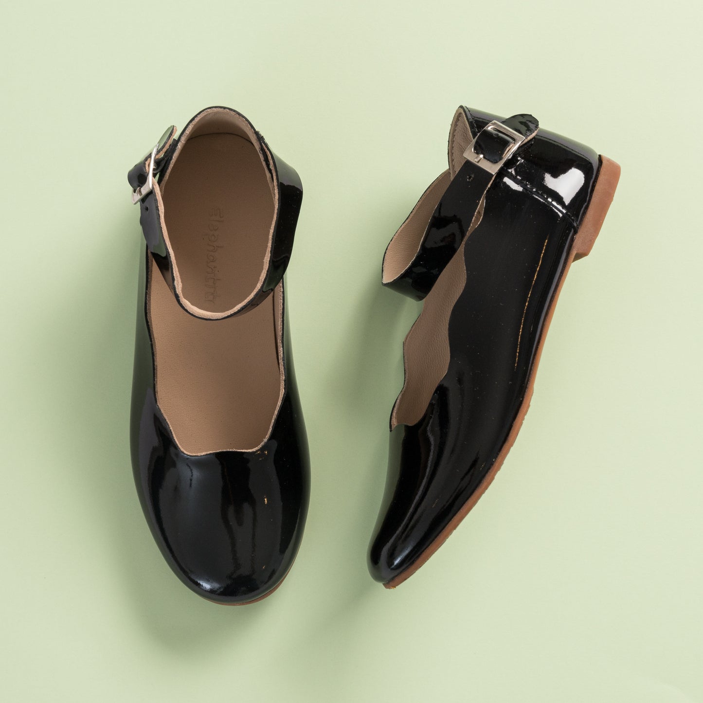 Black patent leather girls' ballet flats with ankle strap and buckle on light green background.