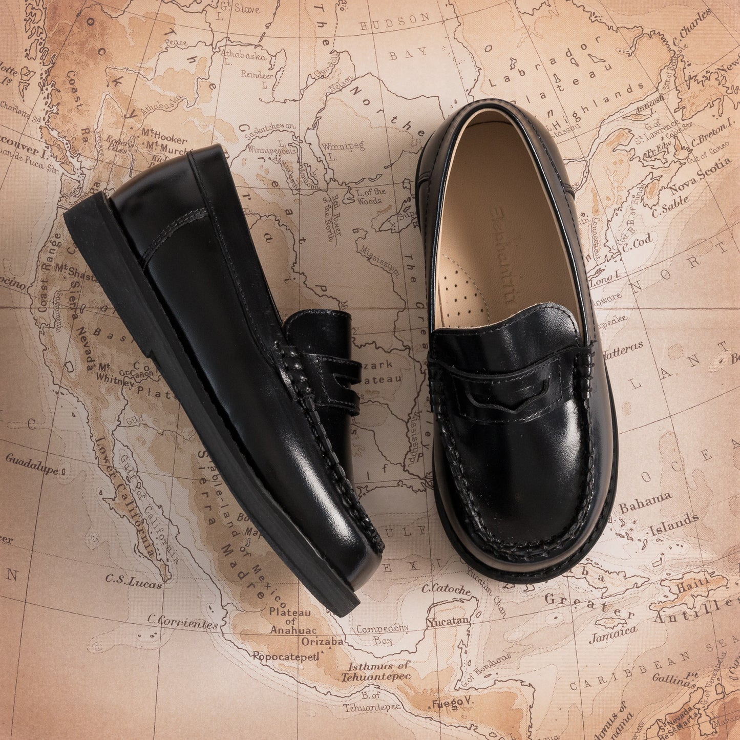 Black glossy leather boys' moccasins with tassel detail and stitched toe on map background.