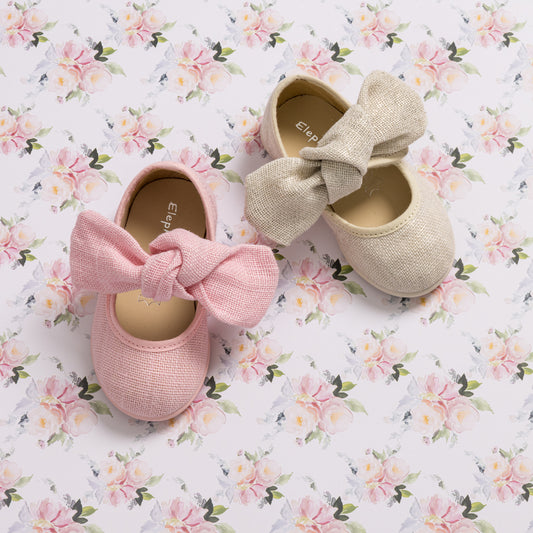 Pair of toddler Mary Jane shoes in pink and beige linen with large bow accents on floral background.