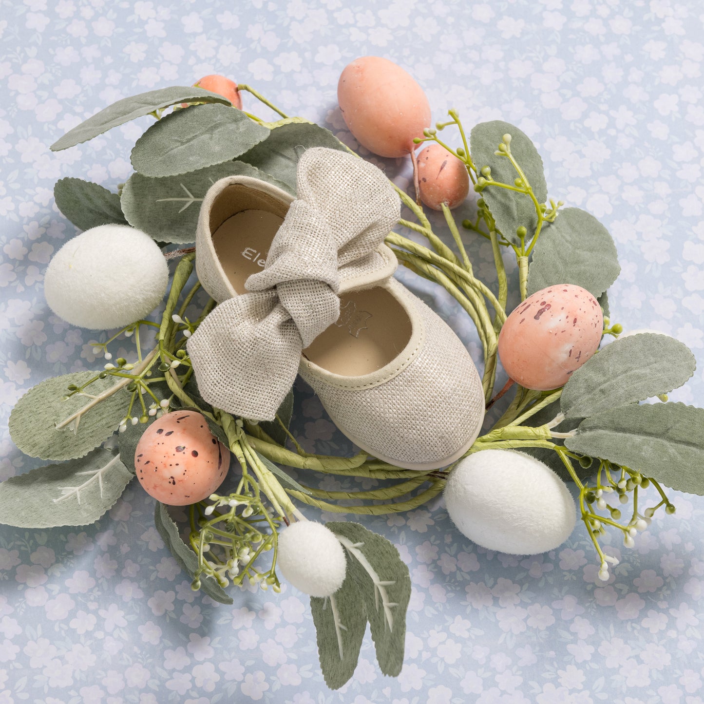 Beige linen toddler Mary Jane shoe with large bow on top, displayed with decorative greenery and pastel Easter eggs.