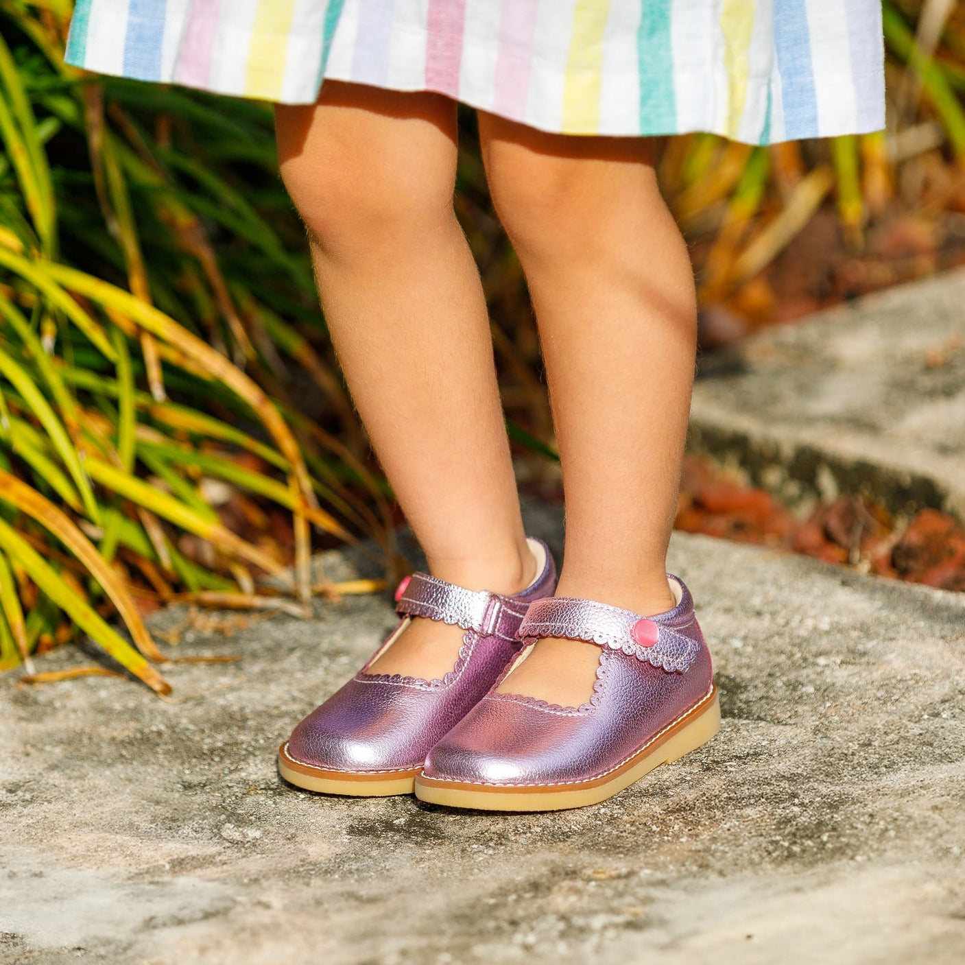 Metallic pink children’s Mary Jane shoes with T-strap and tan rubber sole on toddler feet outdoors.