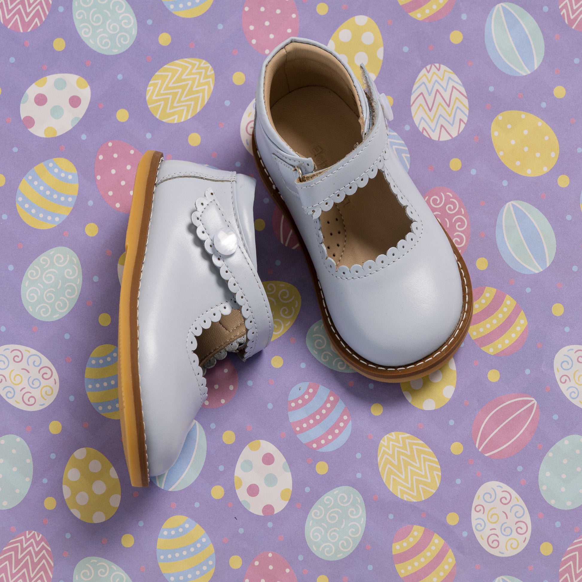 Light blue toddler Mary Jane shoes with scalloped edges and buckle straps on Easter egg patterned background.