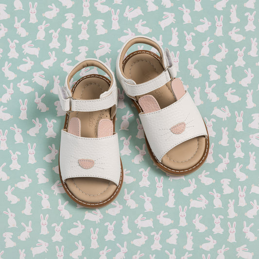 White toddler sandals with bunny cutout detail and adjustable ankle straps on bunny-patterned background.