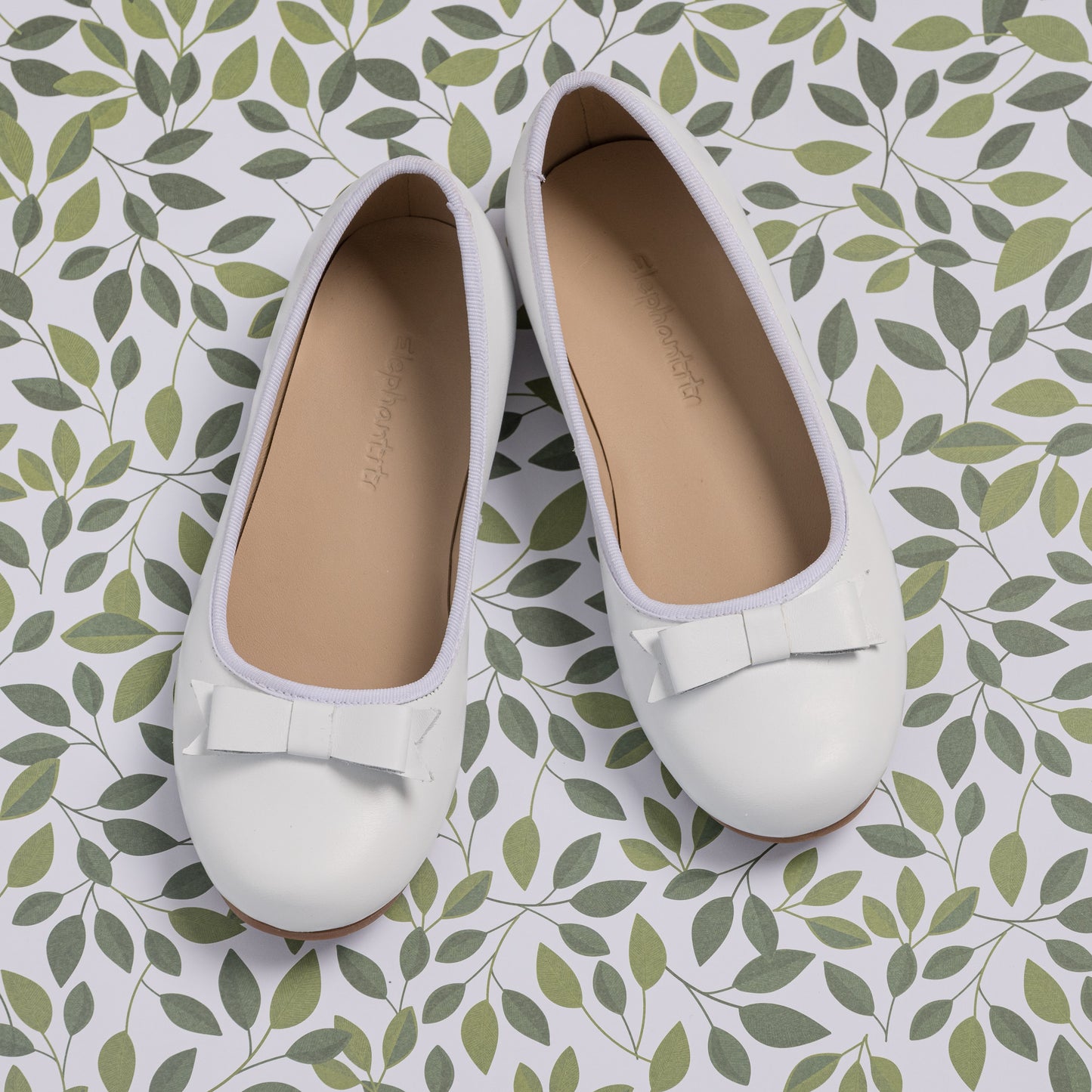 White toddler ballet flats with rounded toes and small decorative bows on top, set on leafy patterned surface.
