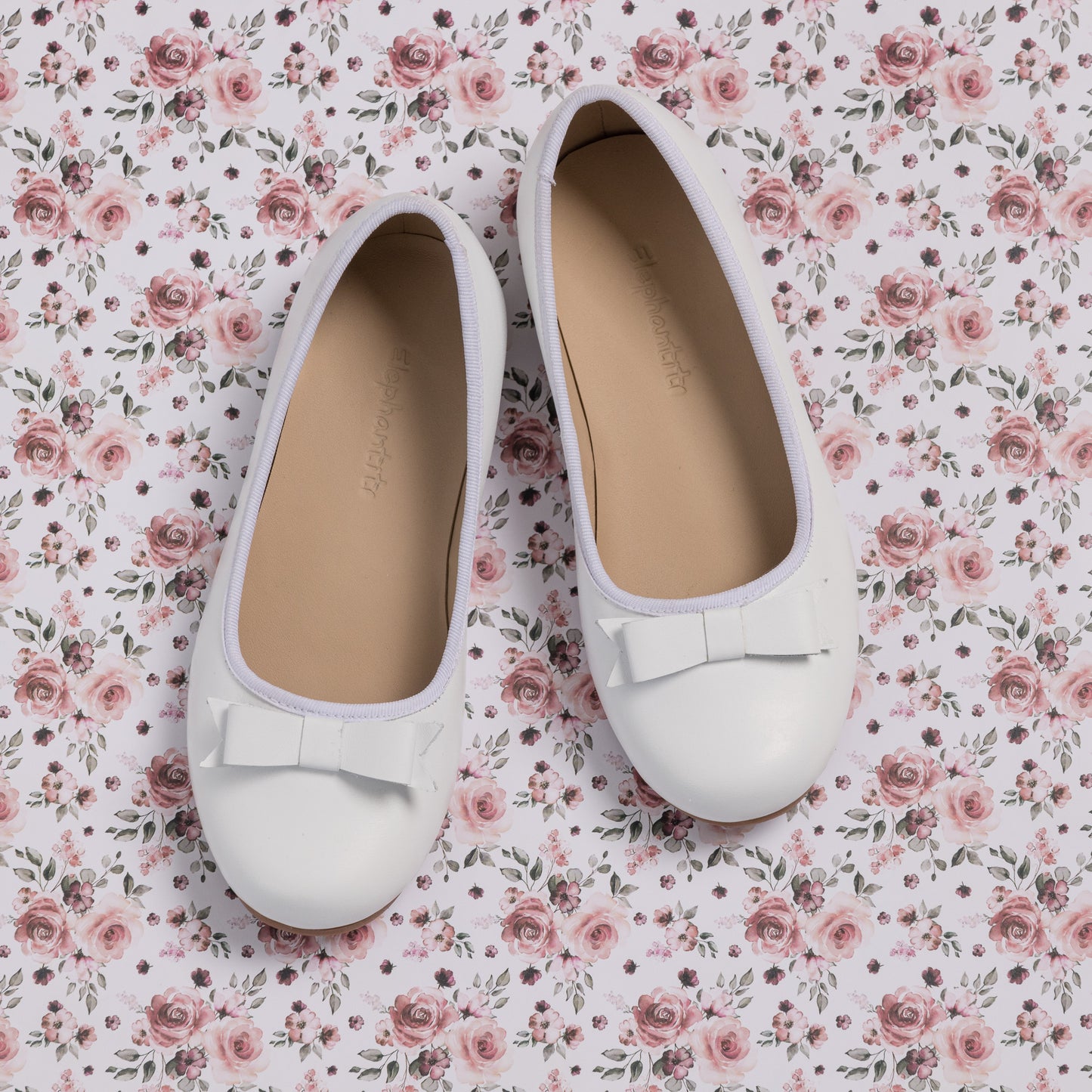 White toddler ballet flats with rounded toes and decorative bows on top, set on floral fabric background.
