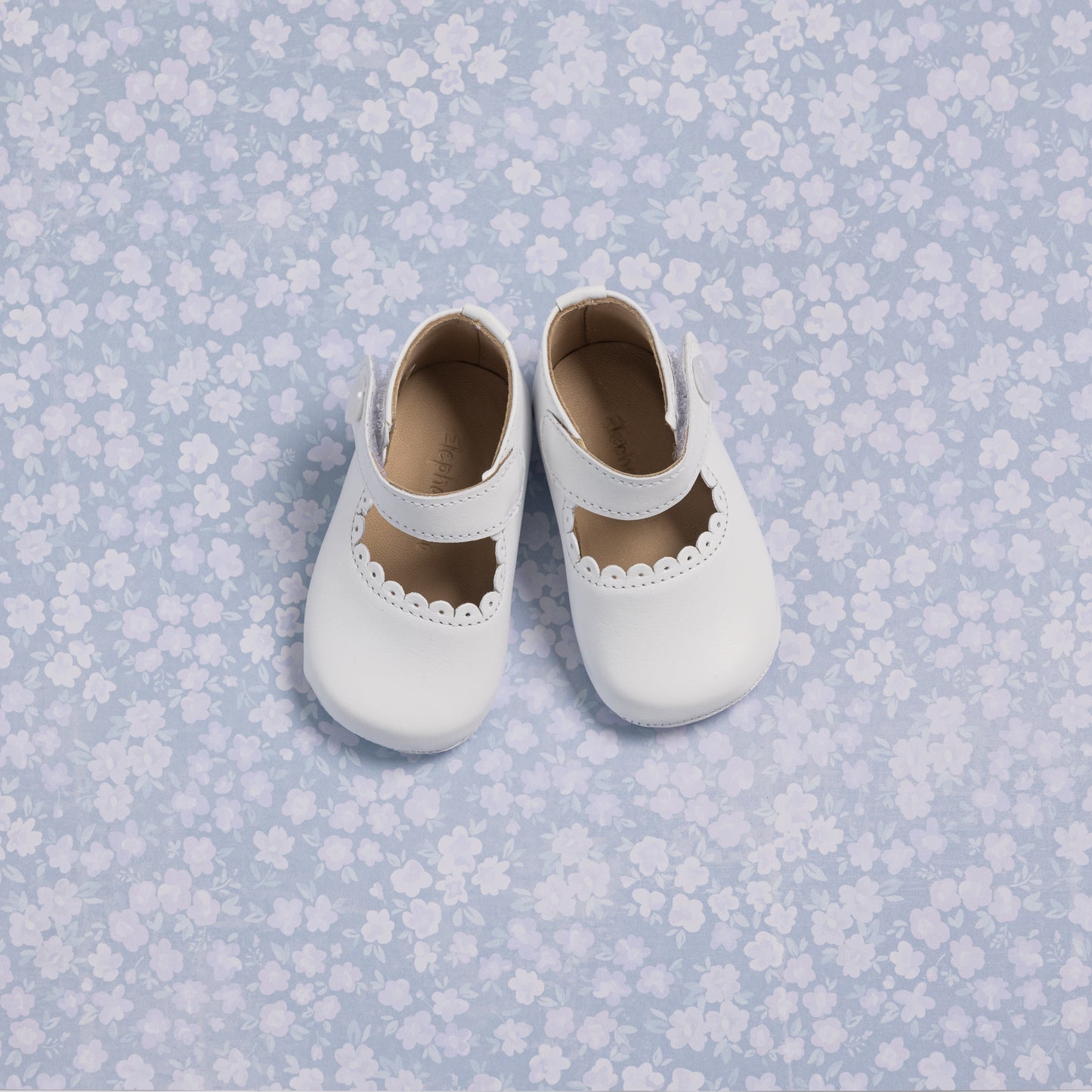 White baby Mary Jane shoes with scalloped edges and a single strap on a blue floral background.