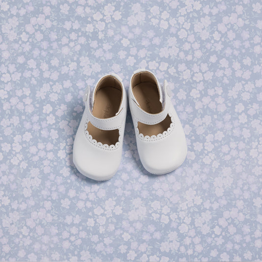 White baby Mary Jane shoes with scalloped edges and a single strap on a blue floral background.