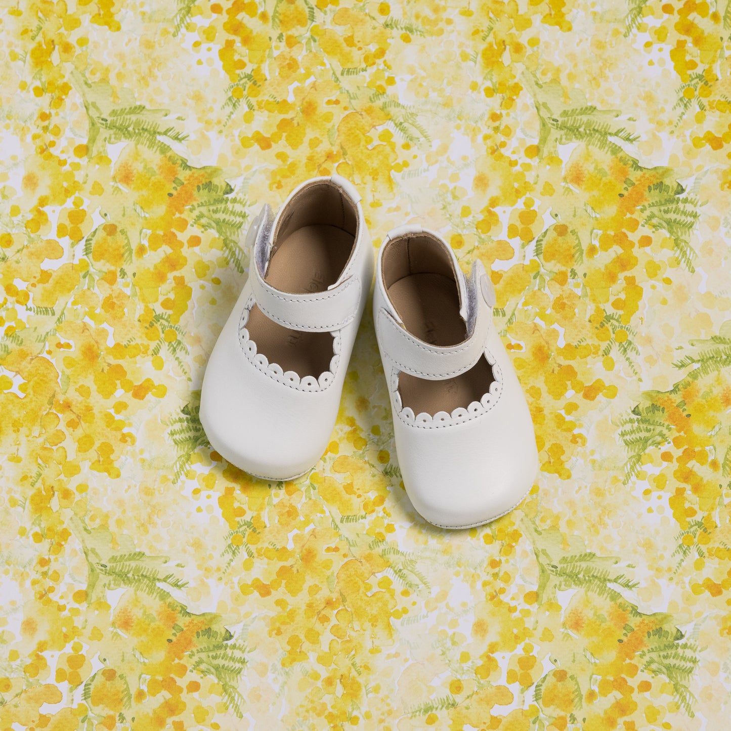 White baby Mary Jane shoes with scalloped edge and strap on yellow floral background.