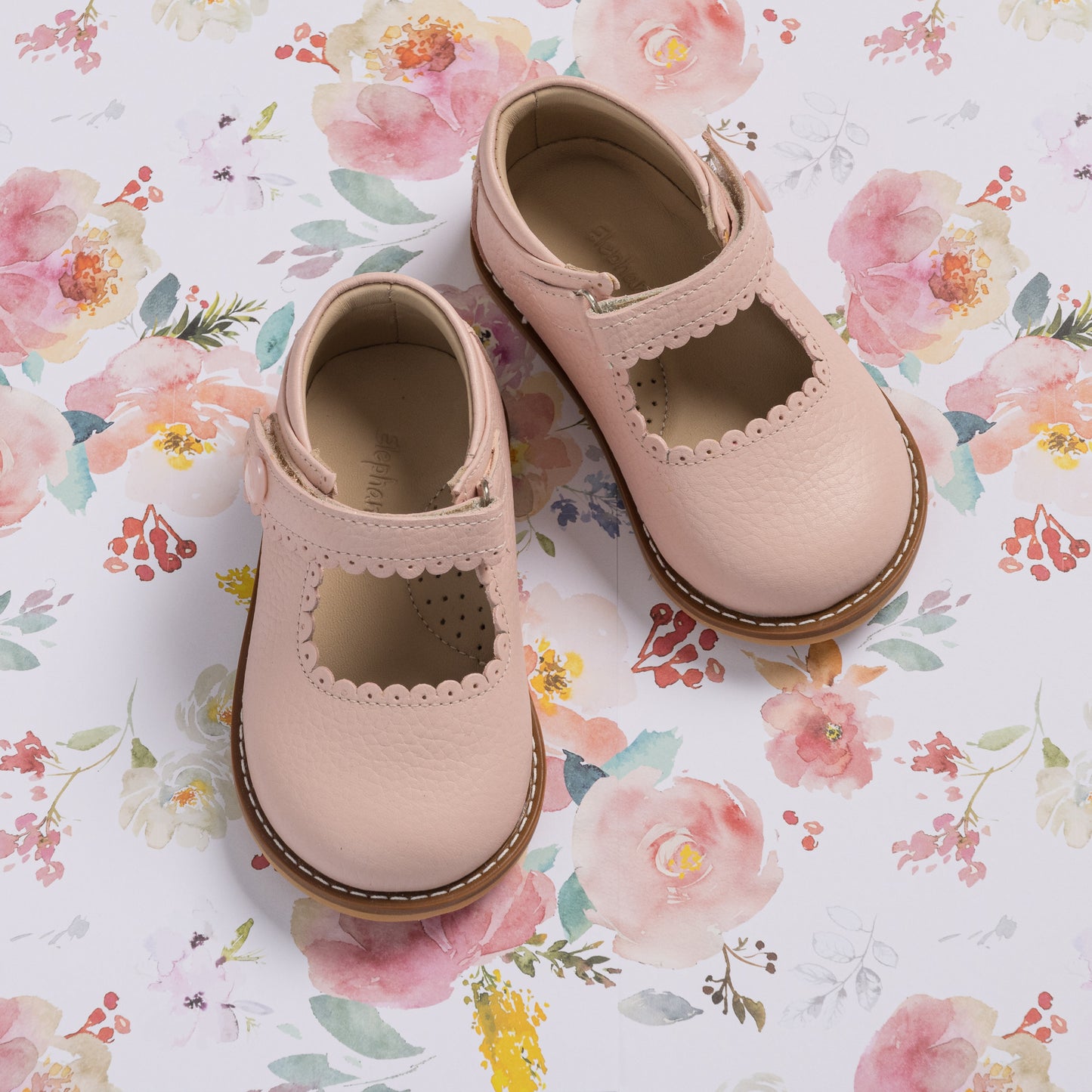 Pink toddler Mary Jane shoes with scalloped edges and brown soles on floral background.