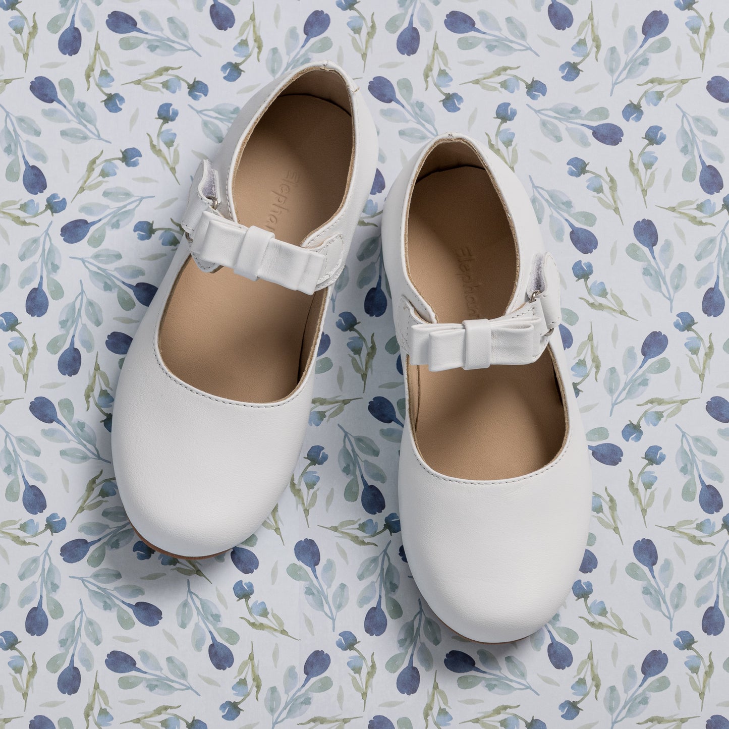 White leather Mary Jane shoes for girls with a buckle strap and rounded toes on a floral background.