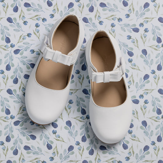 White leather Mary Jane shoes for girls with a buckle strap and rounded toes on a floral background.