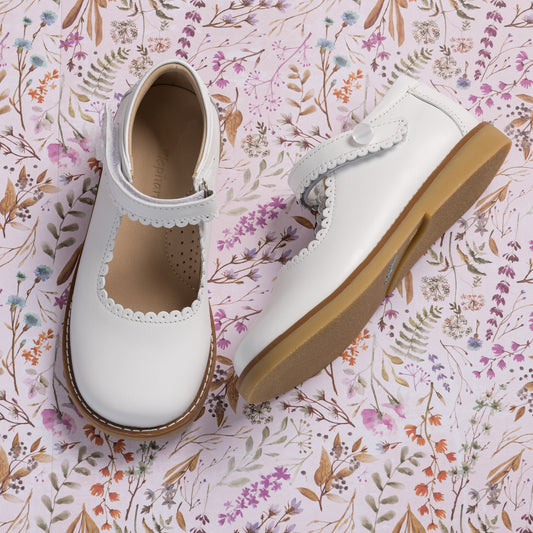 White leather Mary Jane shoes with scalloped edges and velcro strap, tan rubber sole.