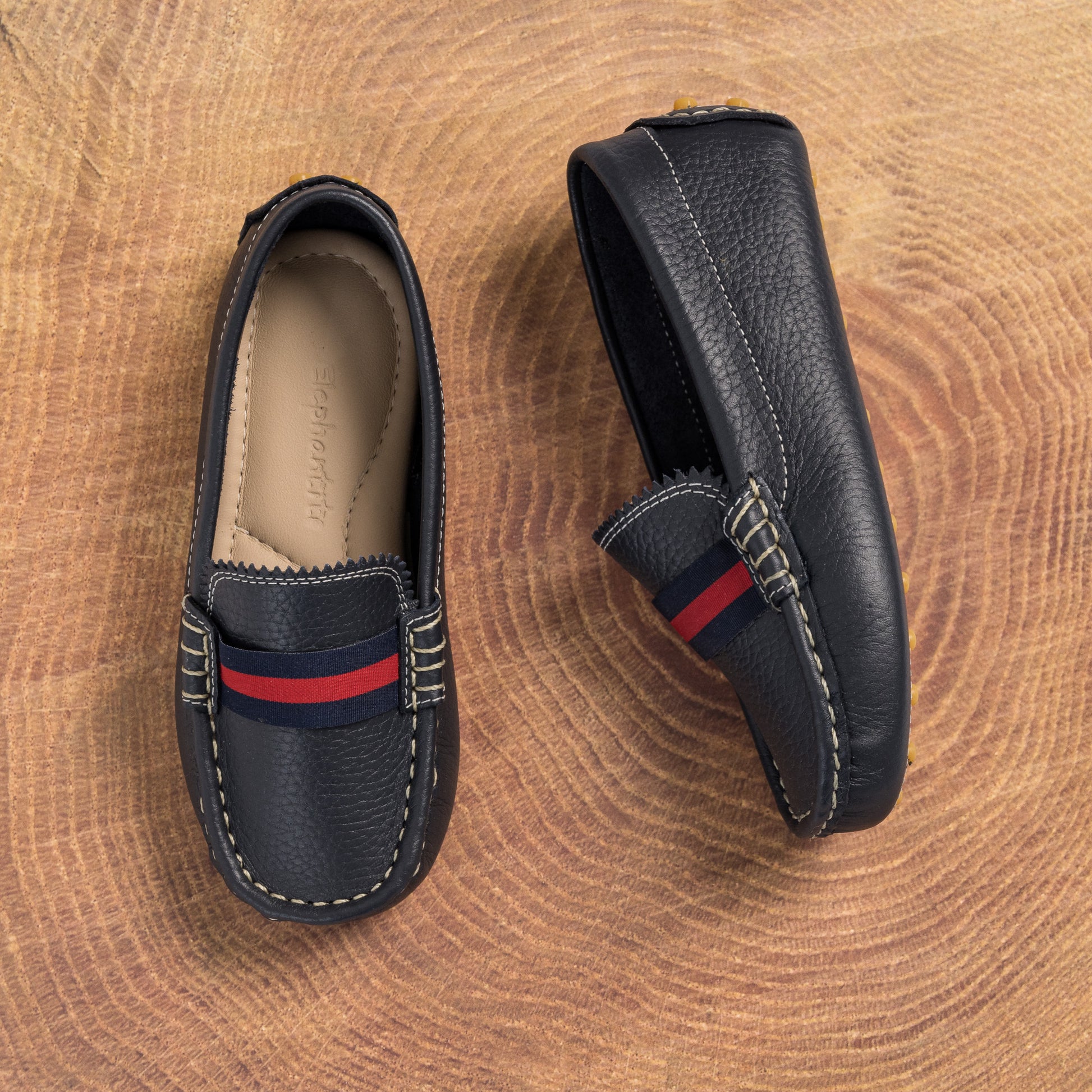 Navy blue leather boys' loafers with red and navy striped strap and white stitching on wooden surface.
