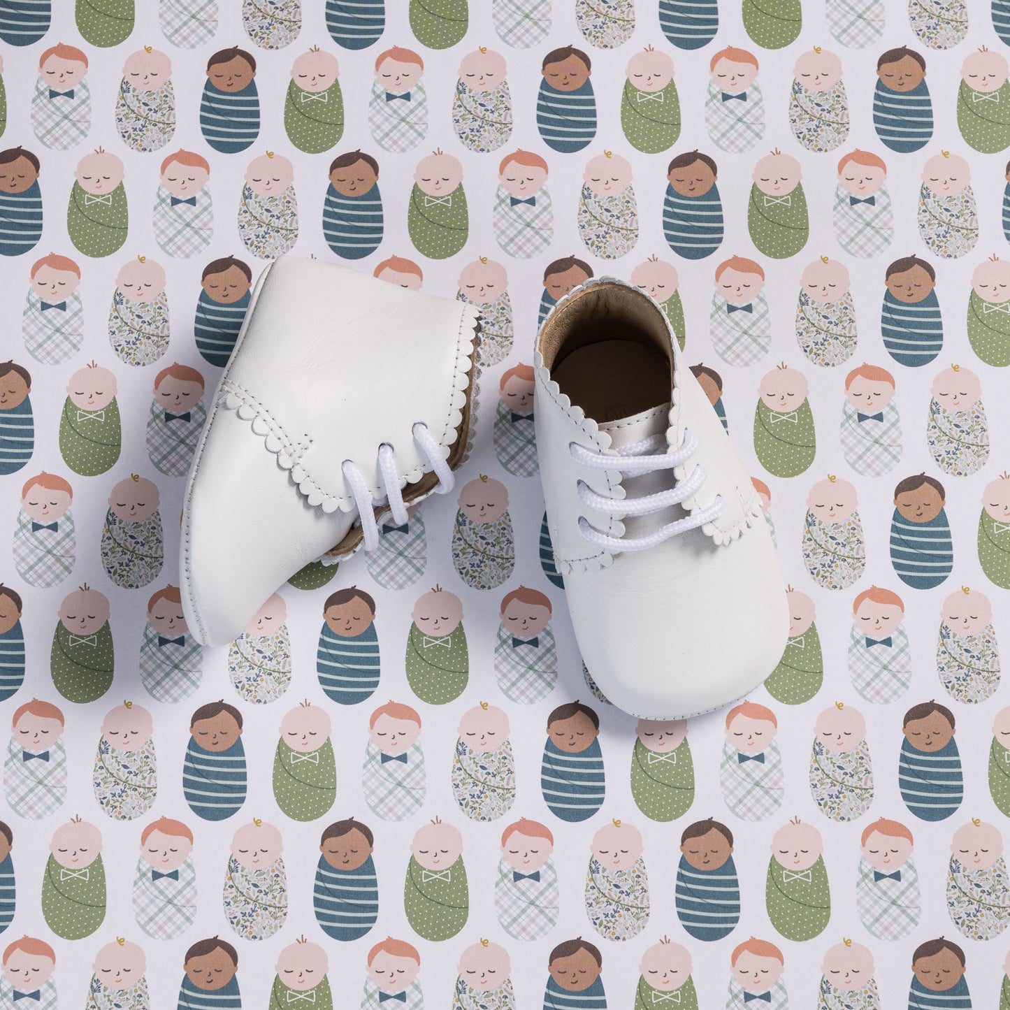 White leather baby booties with scalloped edges and white laces on patterned background.