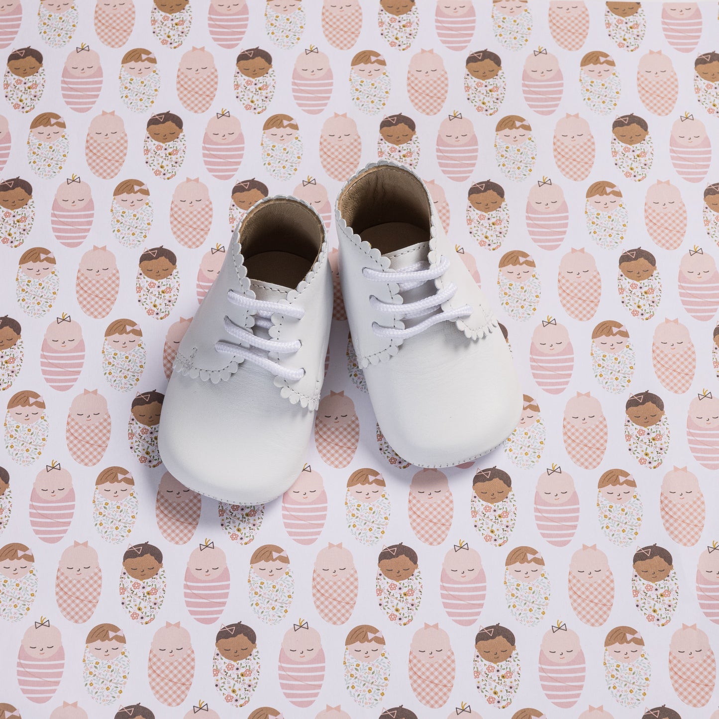White baby booties with scalloped edges and white laces on patterned background.