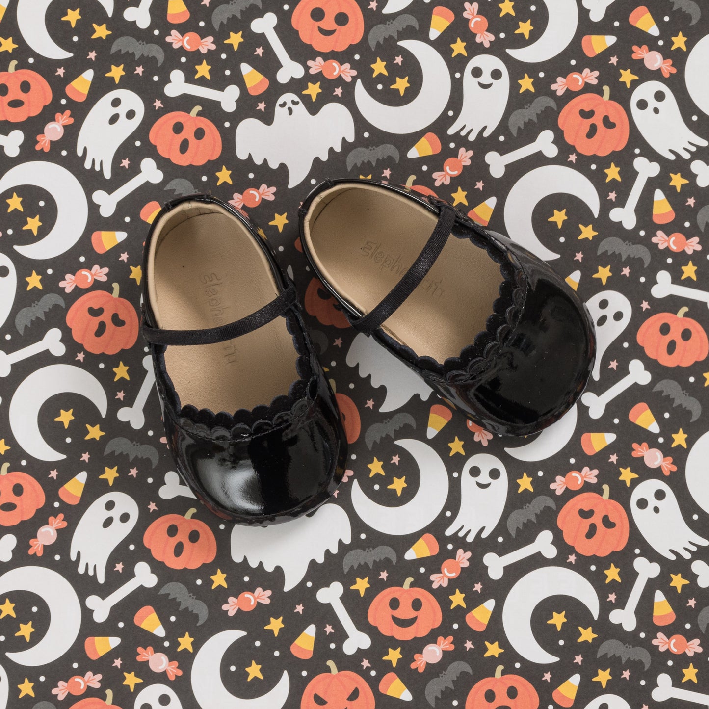 Glossy black patent baby ballet flats with scalloped edges and cross straps on a Halloween-themed background.