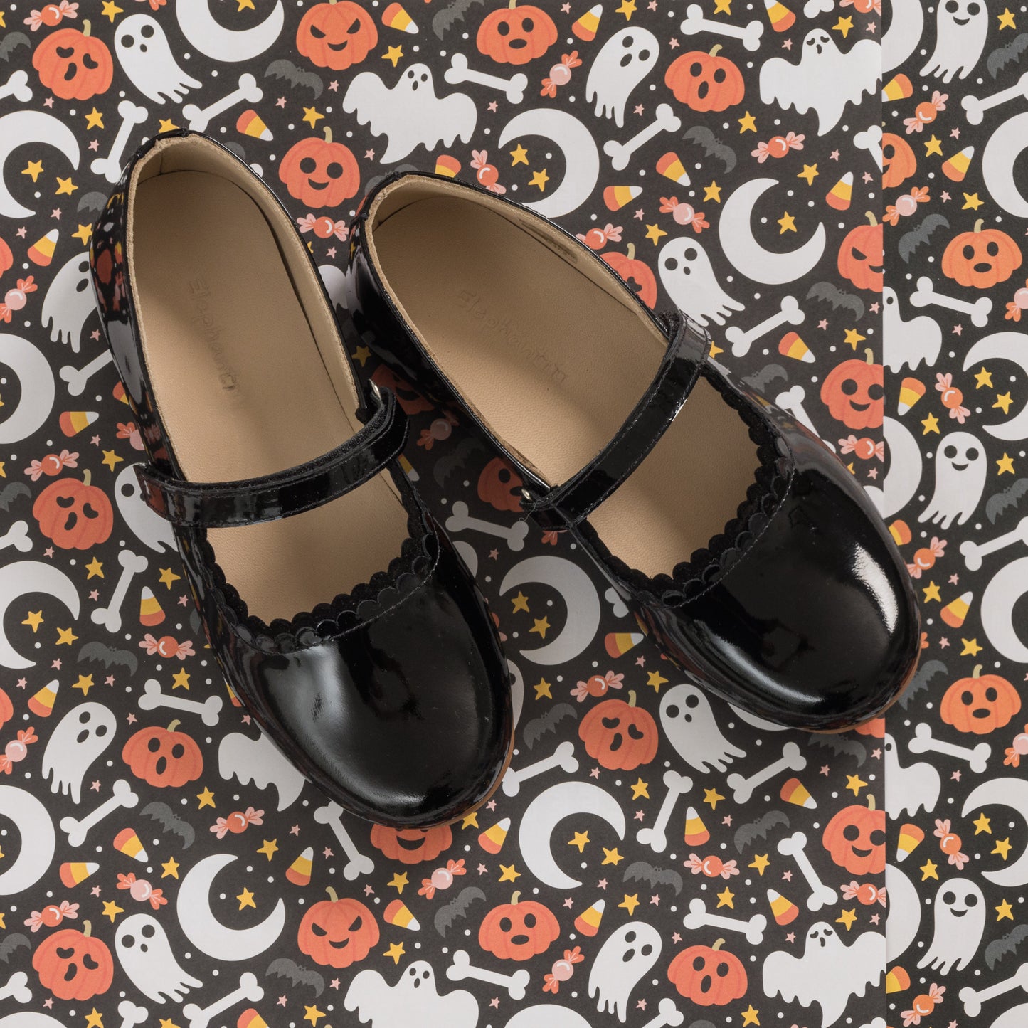 Black patent leather girls' Mary Jane flats with scalloped edges and a single strap buckle on a Halloween-themed background.
